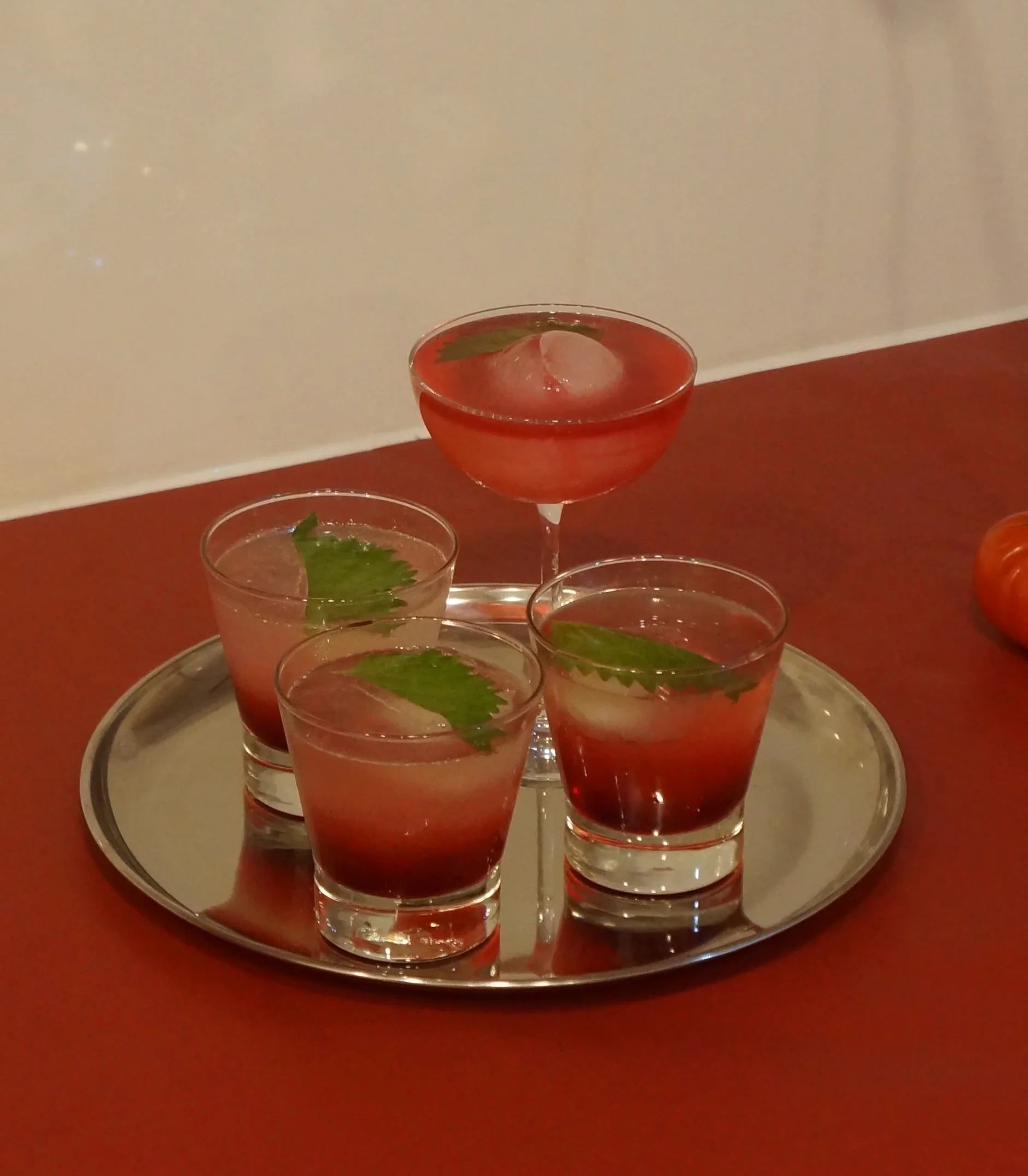 Four Umeboshi Yuzu cocktails on a silver tray on a red table, garnished with shiso leaves and round ice
