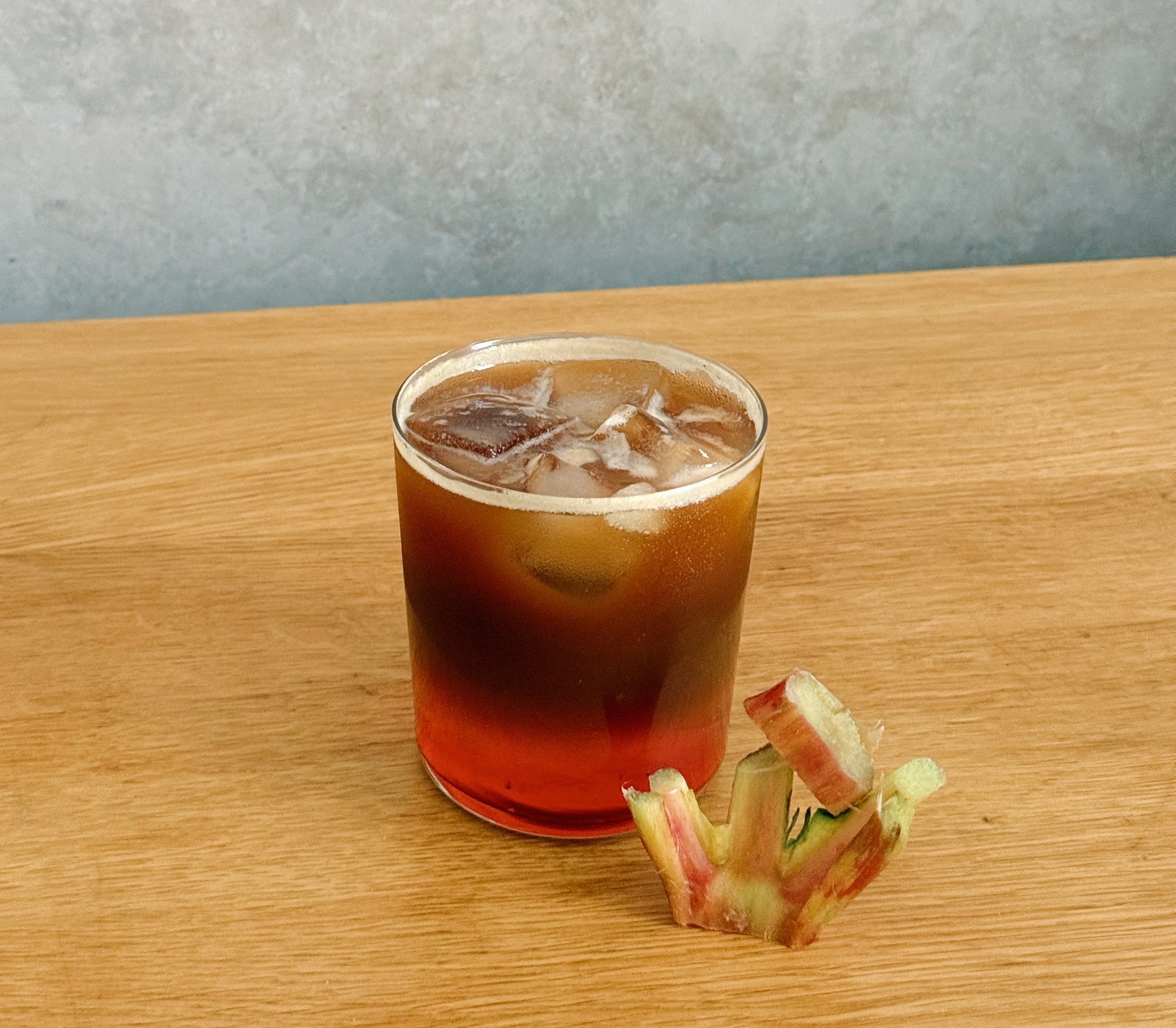 Rhubarb espresso tonic served over ice in a clear lowball glass on a wooden table with a piece of rhubarb