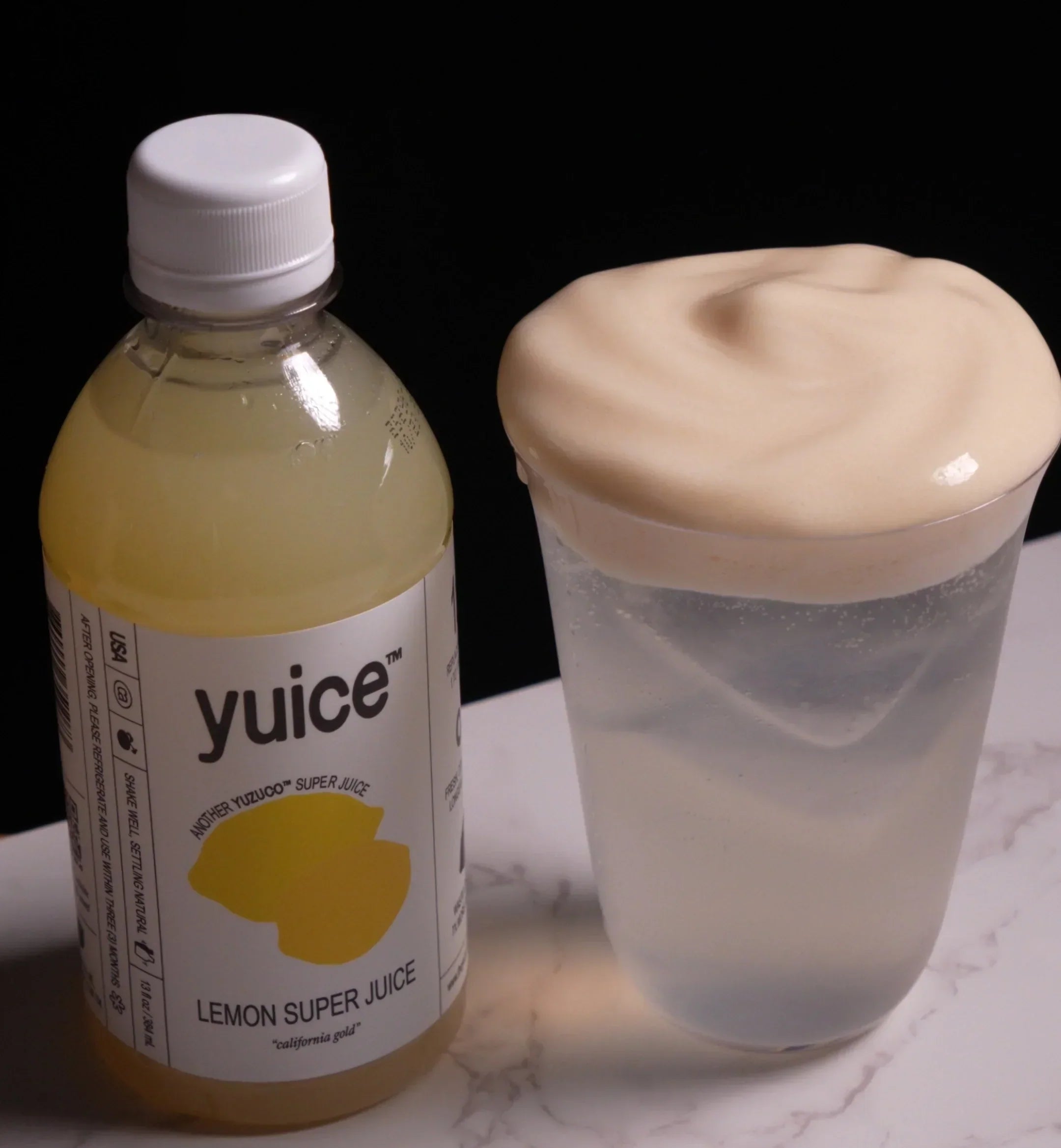 Bottle of Yuice Lemon Super Juice beside a clear cup of fizzy Arnold Palmer lemon sour topped with creamy foam on a marble countertop against a black background
