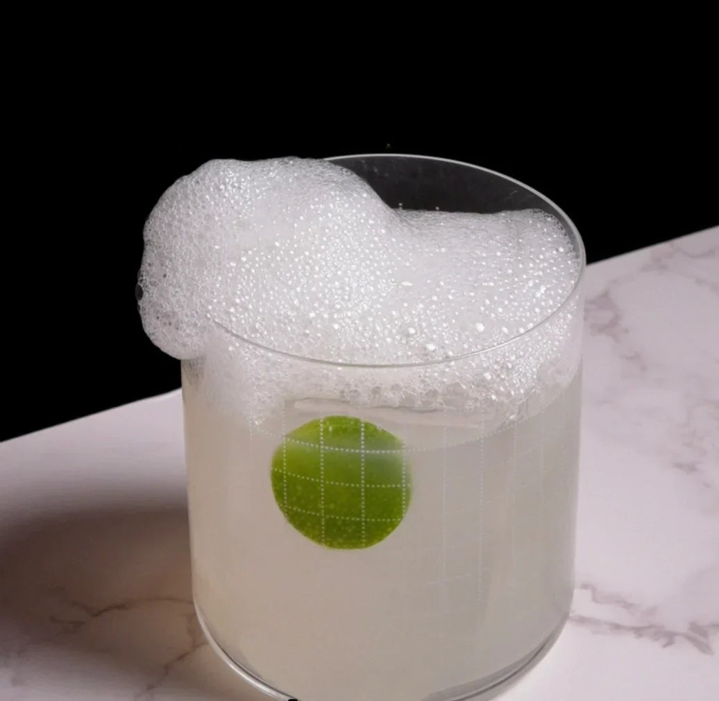 Foamy lime margarita in a short clear glass with a lime wheel, foam spilling over onto a marble surface