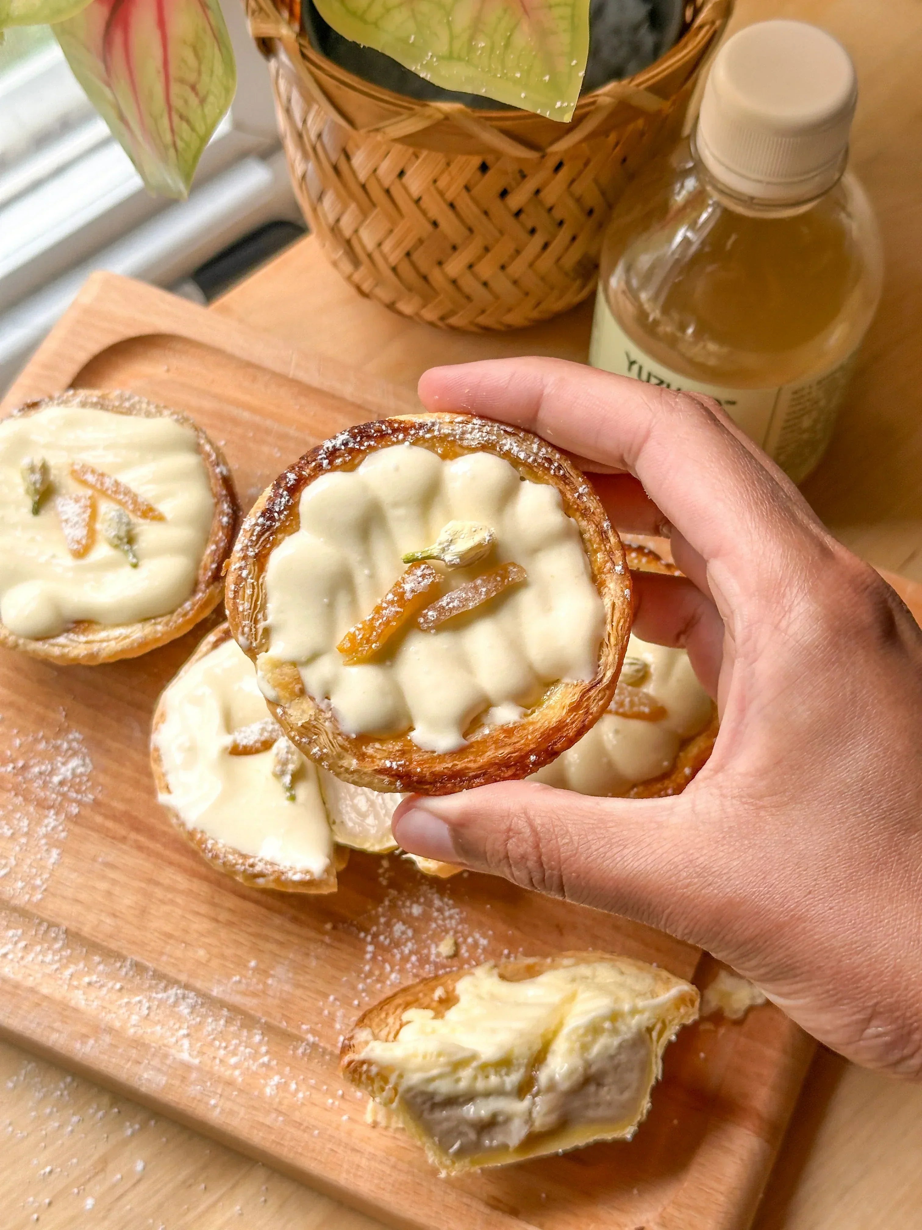 Hand holding a yuzu jasmine egg tart above a wooden board of tarts with powdered sugar and a bottle of yuzu juice