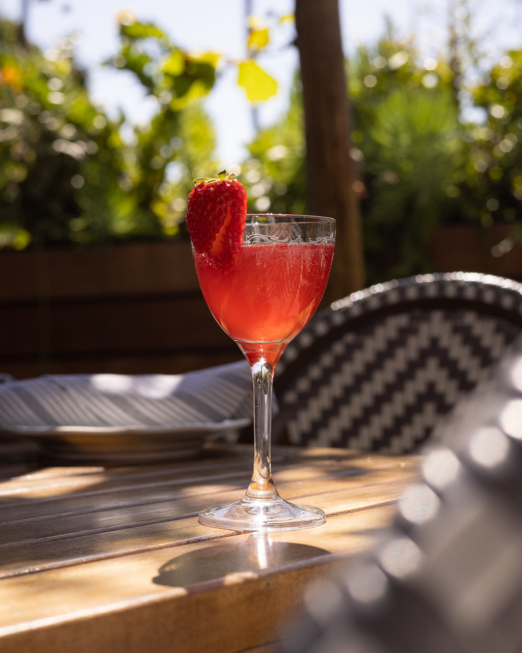 a bright red spring yuzu spritz cocktail with a strawbery garnish in a nick and nora glasssitting on a table on a sunny day