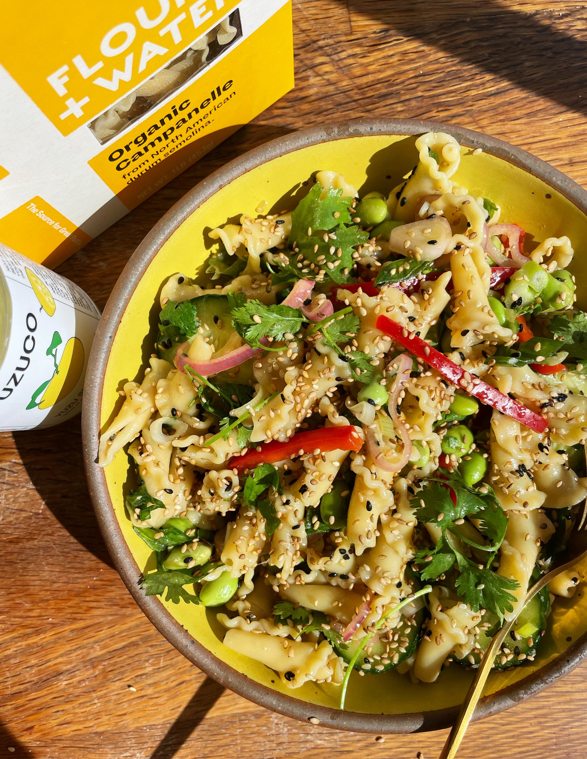 vibrant yellow bowl of campanelle yuzu pasta salad on a wood table. dish sits in front of a bottle of yuzuco yuzu super juice and a box of flour + water dried pasta. 