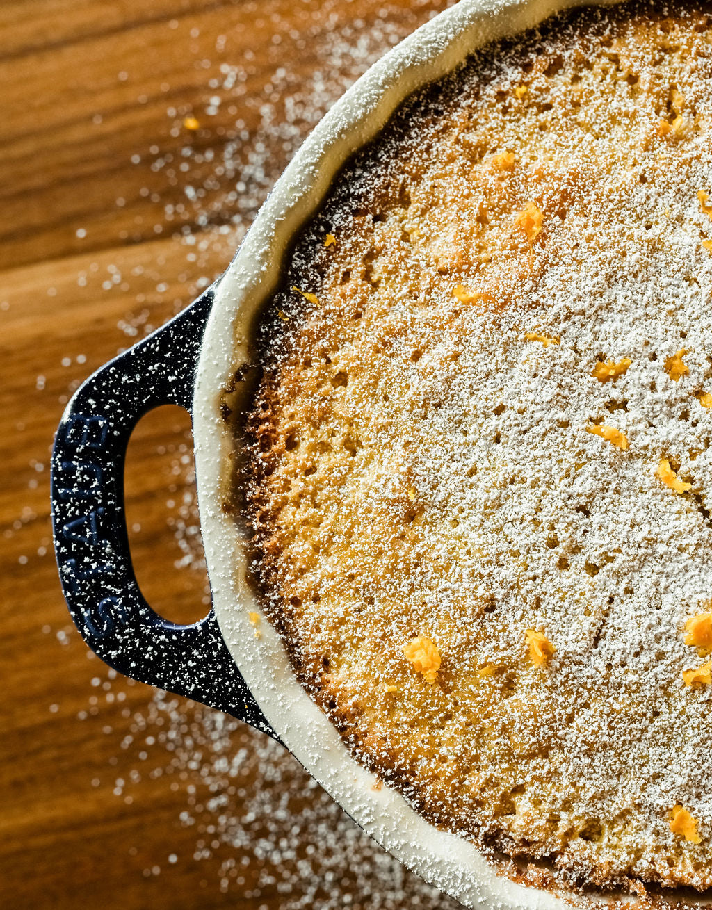 freshly baked yuzuco yuzu olive oil cake covered in yuzu zest in a blue pan on wood table being dusted by powdered sugar