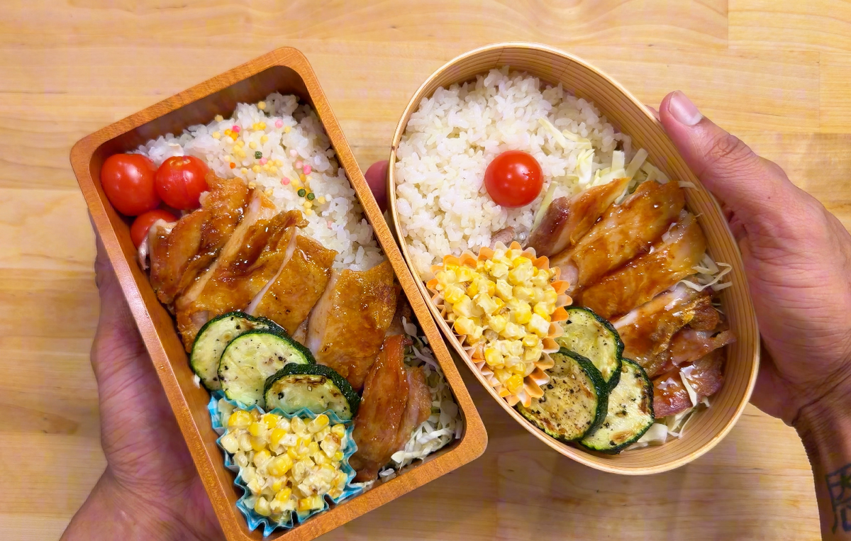 Crispy Yuzu Chicken with Yuzu Kosho Corn, grilled zucchini, tomato, on top of rice. in two bento boxes, left is rectangular, right is oval shaped. held with hands on top of a wooden table top