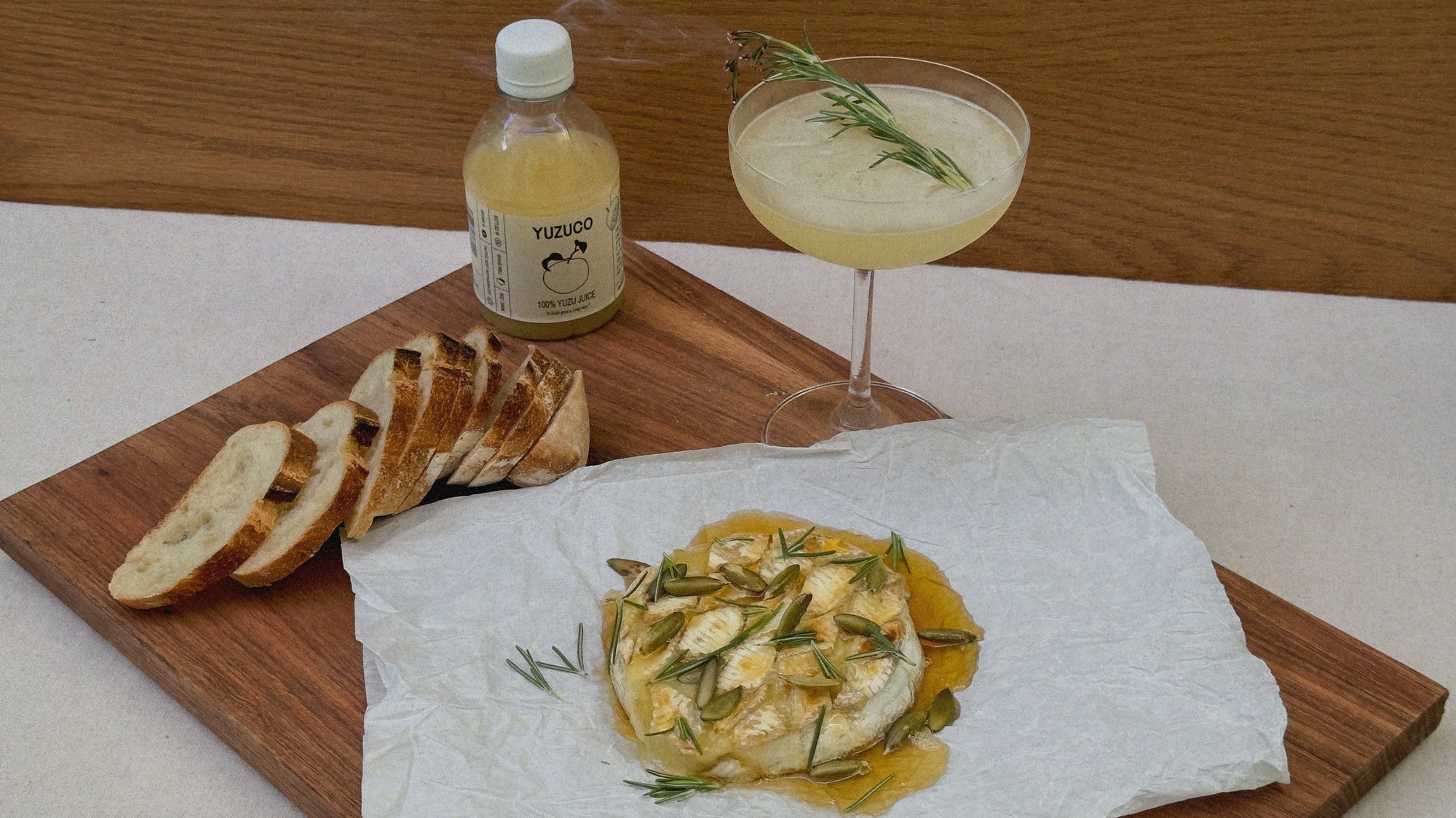 100% yuzu juice from yuzuco next to a sliced baguette a yuzu bees knees cocktial with fresh rosemary and a beautiful yuzu-honey and herb covered baked brie.