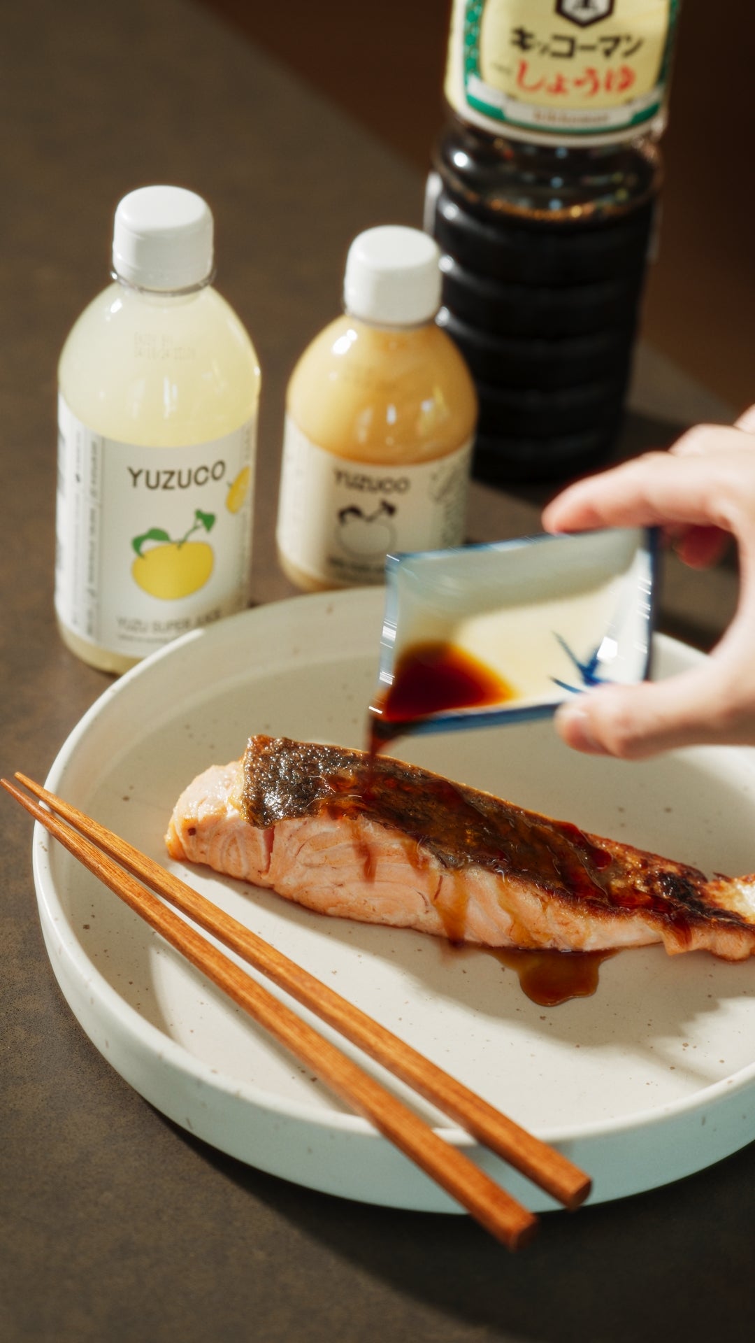 yuzu ponzu being poured over a freshly cooked salmon filet on a plate next to yuzu yuzu juice and yuzu super juice and soy sauce bottles