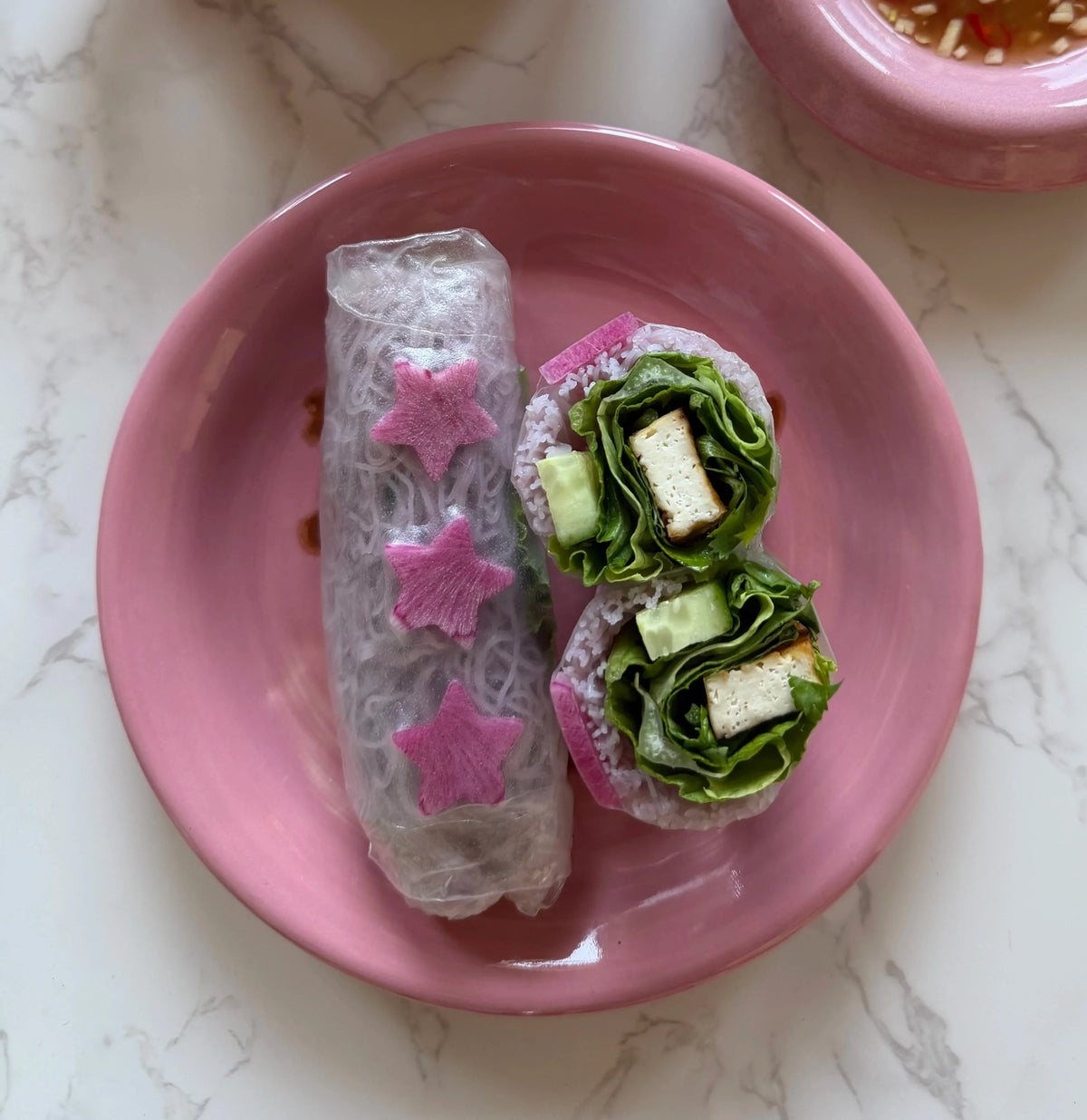 two spring rolls with pink star radishes with one cut open on a pink dish with sauce in the background