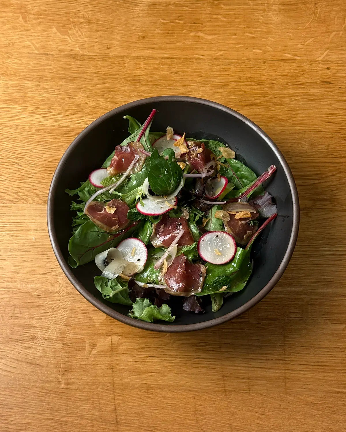 wasabi yuzu salad with greens, sliced radish rounds, duck prosciutto in a black bowl