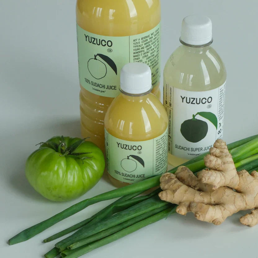 Bottles of Yuzuco Sudachi juice with fresh ginger, green onions, and a green tomato.