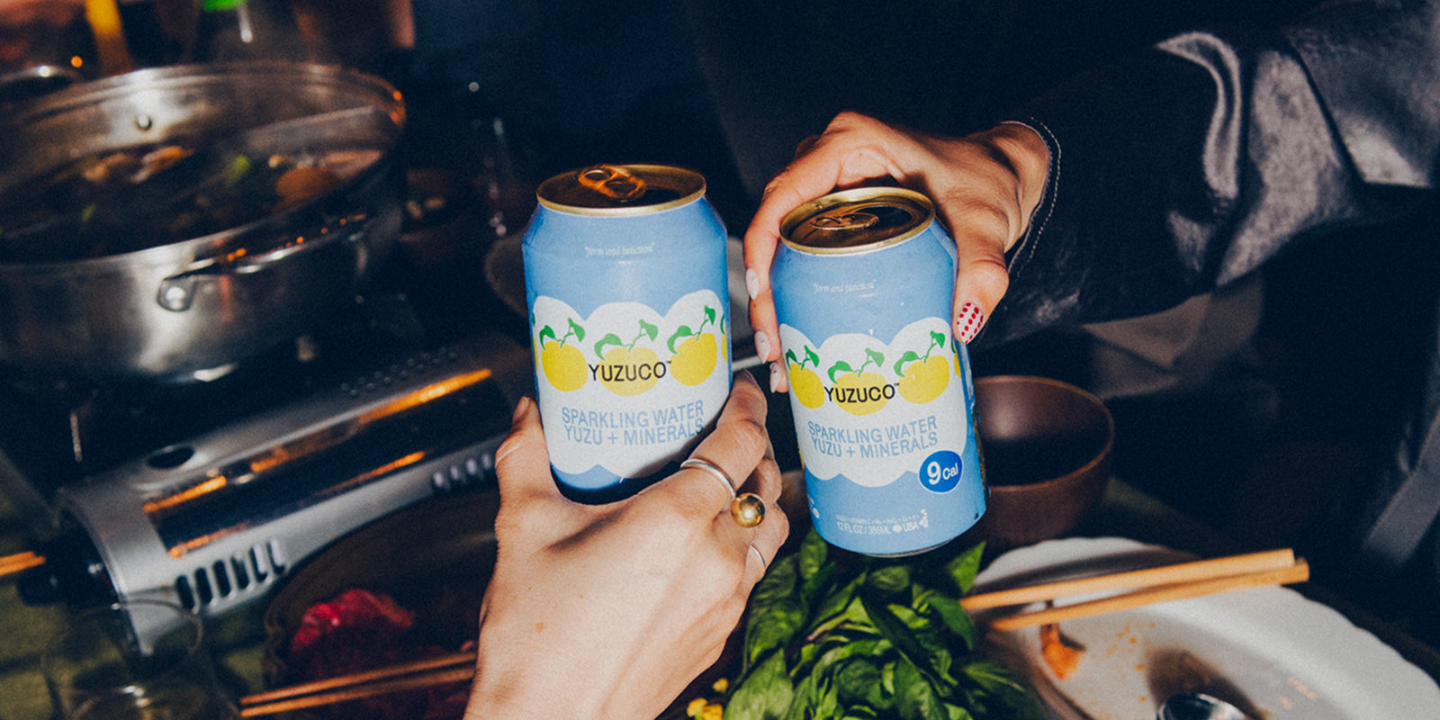 Two hands holding YUZUCO sparkling yuzu cans over a table with hot pot, chopsticks, and shared snacks