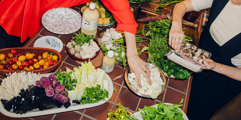 People arranging hot pot ingredients on a tiled table with mushrooms, cherry tomatoes, quail eggs, greens, and YUZUCO bottles