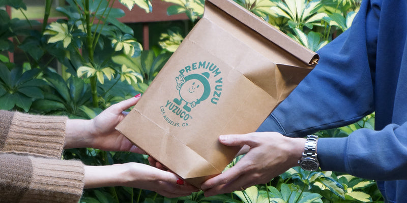 Two people handing a brown paper gift bag stamped with Premium Yuzu logo against green foliage