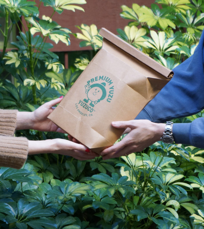 Three-quarter view of Yuzu Gift Bag showcasing green Premium Yuzu logo and hands exchanging it over a leafy outdoor background