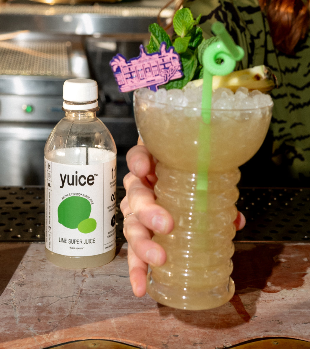 Front view of 13oz Lime Super Juice bottle showcasing the label beside a hand holding a crushed-ice lime cocktail on a bar counter