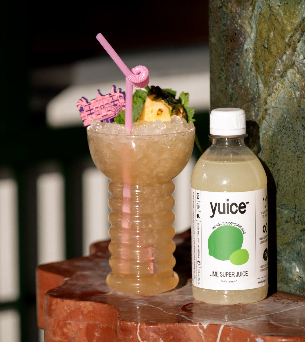 Front view of 13oz Lime Super Juice bottle showcasing white label with green logo and 'yuice' text beside a crushed-ice cocktail on red marble