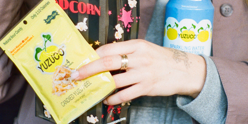 Hand holding YUZUCO candied yuzu peel pouch and YUZUCO sparkling water can on a seated person's lap
