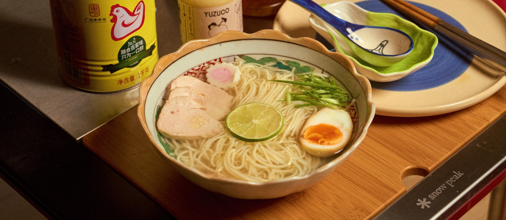 Bowl of ramen topped with a lime slice, soft-boiled egg, sliced chicken, narutomaki, and scallions on a wooden board with chopsticks and ceramic spoons