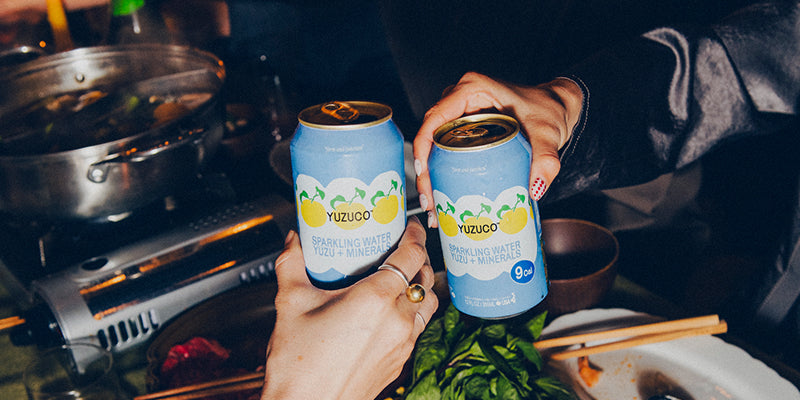 Two hands clinking YUZUCO sparkling water cans over a dimly lit table with hot pot, chopsticks, and greens