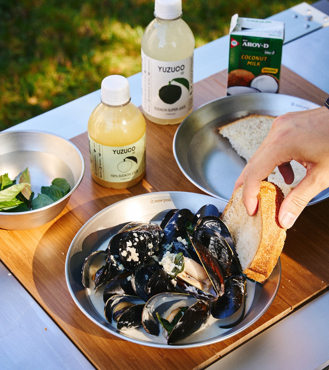 Hand dipping bread into a bowl of mussels on a picnic table beside two Yuzuco Sudachi juice bottles