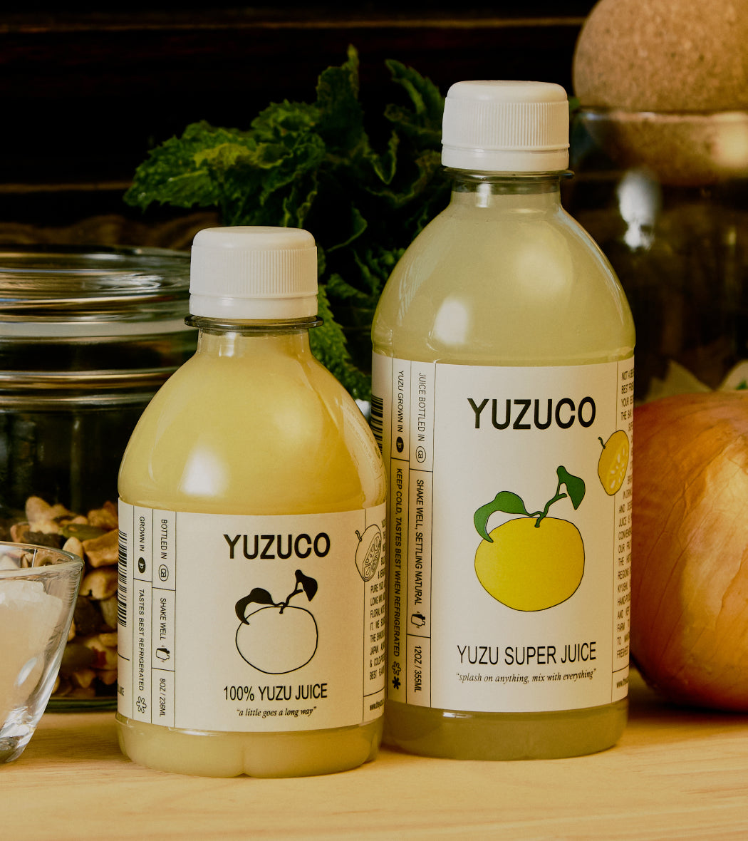 Front view of two YUZUCO juice bottles, small 100 percent Yuzu Juice and larger Yuzu Super Juice on a wooden counter