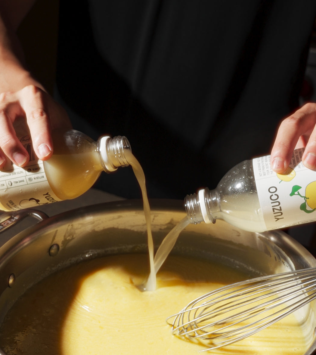 Hands pouring two Yuzu Duo bottles into a saucepan of thick yellow mixture with a whisk inside