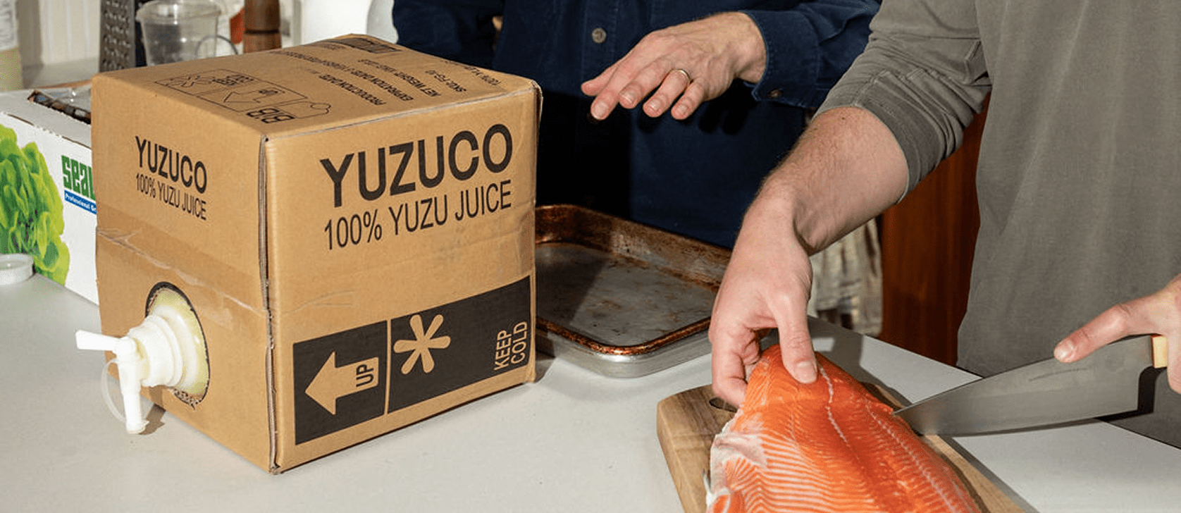 Person cutting a salmon fillet on a cutting board next to a YUZUCO cardboard box labeled 100% yuzu juice in a kitchen
