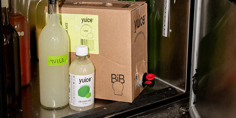 Front view of Yuice Lime Super Juice bottles and cardboard bag-in-box showing tap inside a metal fridge